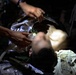 A person is receiving emergency medical treatment in a low-light environment. Medical personnel are attending to the patient, who is lying supine and appears to have an airway management device in place. Various medical supplies are visible around the patient.