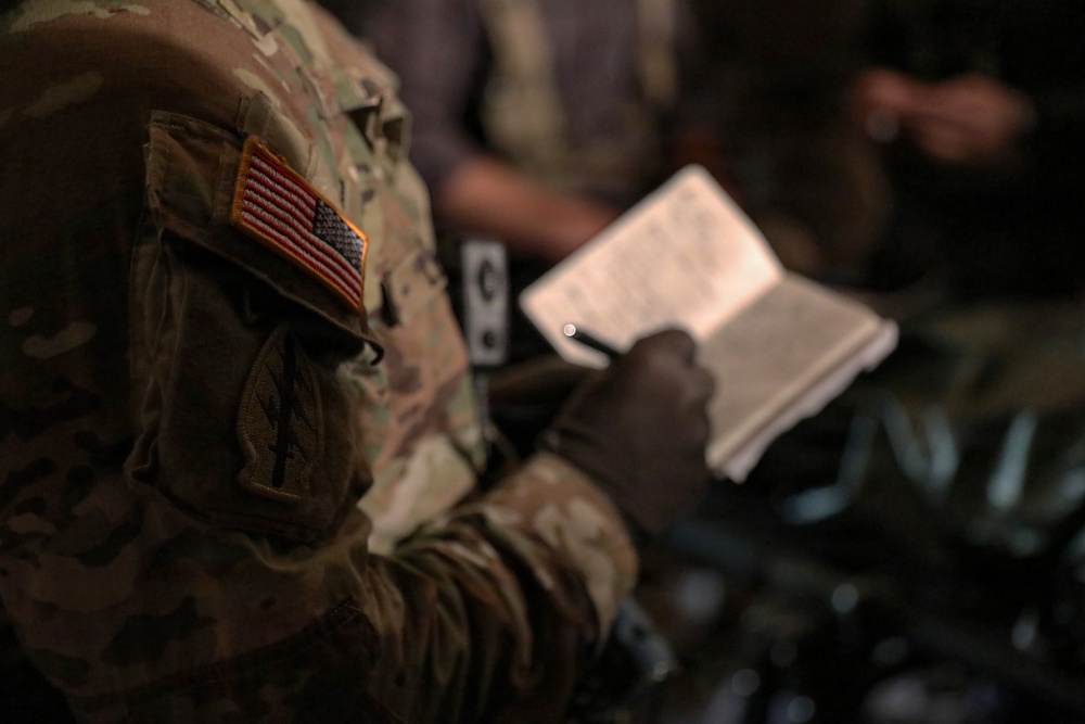 A U.S. military service member in MultiCam/OCP camouflage uniform is writing in a small notebook or notepad. The American flag patch is clearly visible on the right sleeve, displayed in reverse orientation as is standard for U.S. military uniforms. The image is taken in a low-light environment with shallow depth of field, keeping the background blurred.