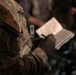 A U.S. military service member in MultiCam/OCP camouflage uniform is writing in a small notebook or notepad. The American flag patch is clearly visible on the right sleeve, displayed in reverse orientation as is standard for U.S. military uniforms. The image is taken in a low-light environment with shallow depth of field, keeping the background blurred.