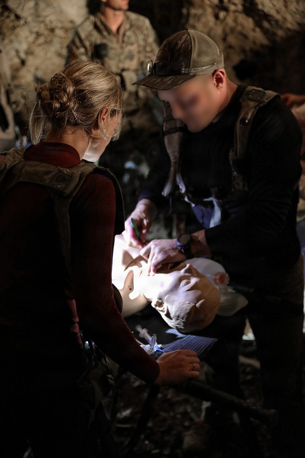 Two individuals in tactical/military gear are performing medical training on a CPR/medical mannequin in what appears to be a cave or rocky environment. One person wears a baseball cap and dark clothing with a tactical vest, while the other has blonde hair in a bun and wears a dark red top with a tactical vest. Additional personnel in military uniforms are visible in the background observing the exercise.