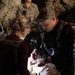 Two individuals in tactical/military gear are performing medical training on a CPR/medical mannequin in what appears to be a cave or rocky environment. One person wears a baseball cap and dark clothing with a tactical vest, while the other has blonde hair in a bun and wears a dark red top with a tactical vest. Additional personnel in military uniforms are visible in the background observing the exercise.