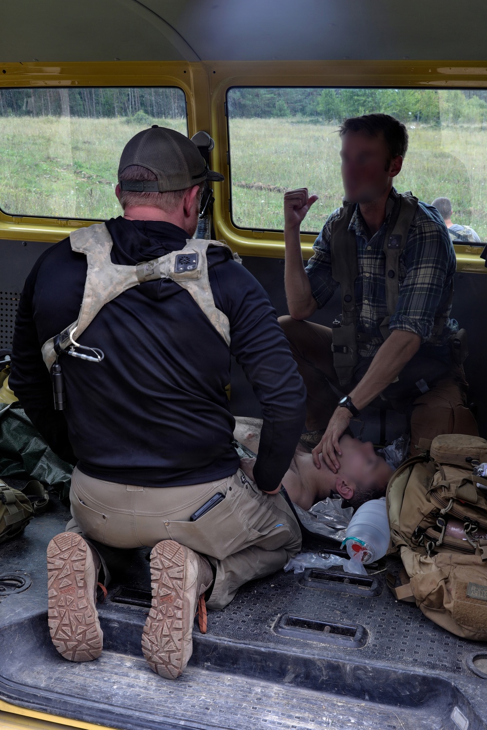 The image shows what appears to be a tactical medical or casualty care scenario inside the cargo area of a yellow vehicle, possibly a van or bus. Two standing/kneeling individuals are attending to a third person who is lying on the floor of the vehicle. Medical supplies and tactical gear are visible around them.
