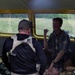 The image shows what appears to be a tactical medical or casualty care scenario inside the cargo area of a yellow vehicle, possibly a van or bus. Two standing/kneeling individuals are attending to a third person who is lying on the floor of the vehicle. Medical supplies and tactical gear are visible around them.