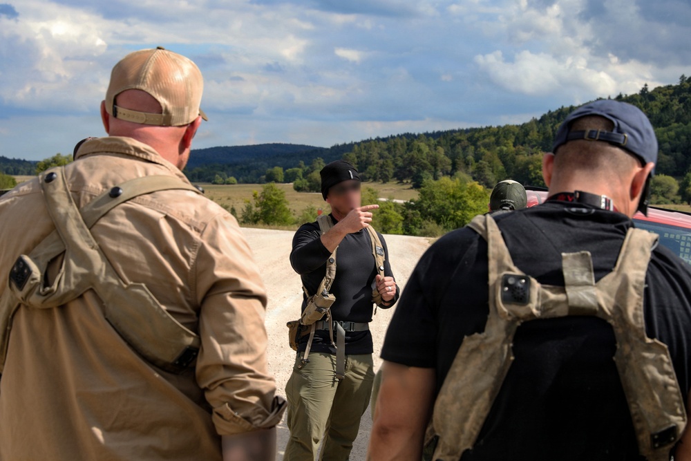 Three individuals in tactical/outdoor gear are gathered outdoors on an elevated position overlooking a forested valley. The central figure, whose face has been deliberately blurred, appears to be pointing and briefing the other two. The setting and equipment suggest a tactical training, reconnaissance, or operational planning scenario.