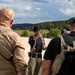 Three individuals in tactical/outdoor gear are gathered outdoors on an elevated position overlooking a forested valley. The central figure, whose face has been deliberately blurred, appears to be pointing and briefing the other two. The setting and equipment suggest a tactical training, reconnaissance, or operational planning scenario.