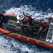 An aerial view of a U.S. Coast Guard rigid-hull inflatable boat (RHIB) underway on open ocean waters. The orange vessel is crewed by multiple personnel in blue uniforms and life vests, and appears to be connected to a larger vessel via a tow line or davit cable. The boat is creating a visible wake, indicating active movement through choppy blue water.