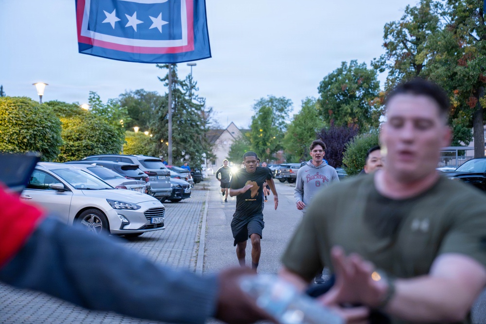 A group of people are running along a paved path in what appears to be an early morning or evening setting, likely on a military installation or organized community event. A blue flag with stars and a red/white border is visible in the upper left corner, suggesting a military or organizational affiliation. Parked cars line the left side of the path, and trees are visible in the background.