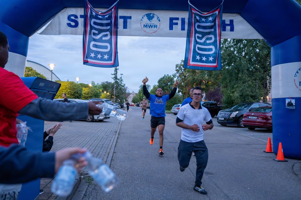 A running race is taking place under an inflatable START/FINISH arch branded with USO and U.S. Army MWR logos. Runners are crossing or approaching the finish line, with one runner in a blue USO shirt celebrating with raised arms. Volunteers or staff on the sides are handing out water bottles to finishers.