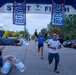 A running race is taking place under an inflatable START/FINISH arch branded with USO and U.S. Army MWR logos. Runners are crossing or approaching the finish line, with one runner in a blue USO shirt celebrating with raised arms. Volunteers or staff on the sides are handing out water bottles to finishers.