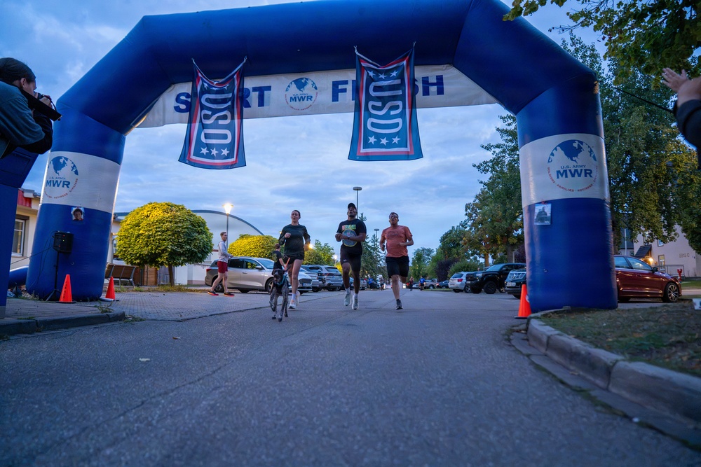 Three runners and a dog are crossing under an inflatable START/FINISH arch at what appears to be a military community fun run organized by USO and U.S. Army MWR (Morale, Welfare and Recreation). The event is taking place at dusk or early morning in what appears to be a military installation, with spectators and a photographer visible on the sidelines. The arch prominently displays USO banners and MWR branding.