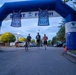 Three runners and a dog are crossing under an inflatable START/FINISH arch at what appears to be a military community fun run organized by USO and U.S. Army MWR (Morale, Welfare and Recreation). The event is taking place at dusk or early morning in what appears to be a military installation, with spectators and a photographer visible on the sidelines. The arch prominently displays USO banners and MWR branding.