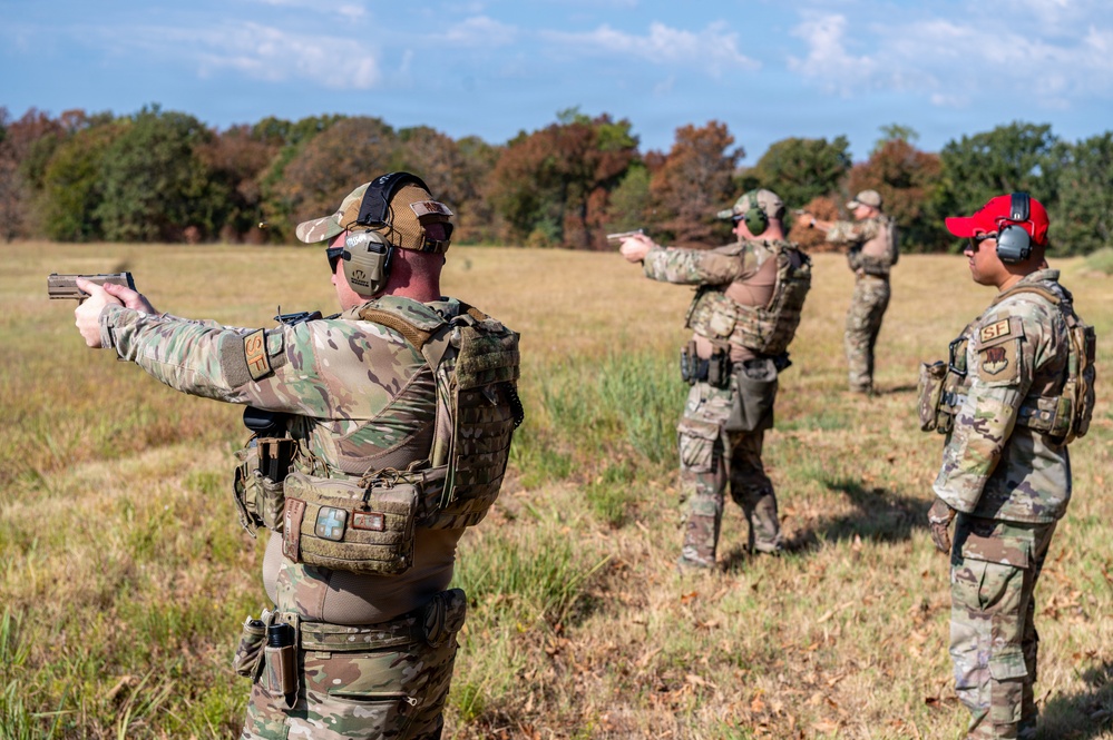 A group of military or law enforcement personnel are conducting an outdoor pistol shooting exercise or qualification on a grassy range. Multiple individuals in multicam/OCP camouflage uniforms and tactical gear are actively firing semi-automatic pistols downrange. One individual wearing a red cap appears to be observing or instructing the others.