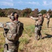 A group of military or law enforcement personnel are conducting an outdoor pistol shooting exercise or qualification on a grassy range. Multiple individuals in multicam/OCP camouflage uniforms and tactical gear are actively firing semi-automatic pistols downrange. One individual wearing a red cap appears to be observing or instructing the others.
