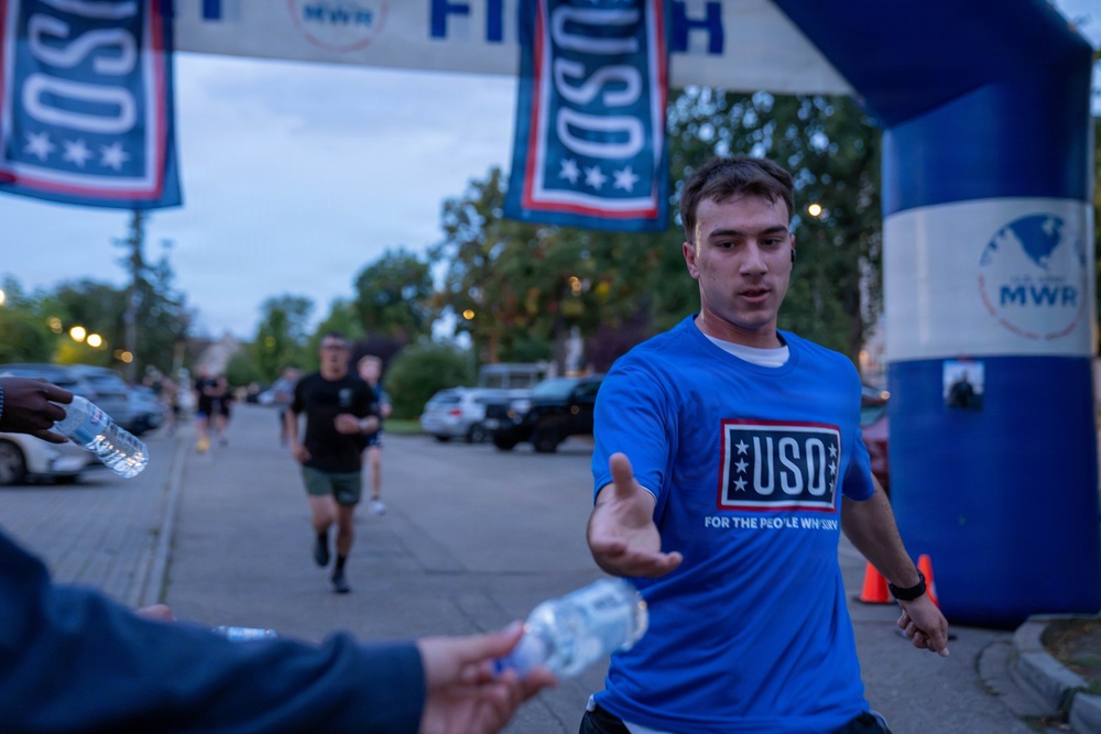 A runner wearing a blue USO t-shirt is crossing or approaching a finish line at what appears to be a charity or military-affiliated 5K/fun run event. He is reaching out to accept a water bottle being handed to him by a volunteer. The event is co-sponsored by USO and MWR (Morale, Welfare, and Recreation), suggesting a military installation setting.