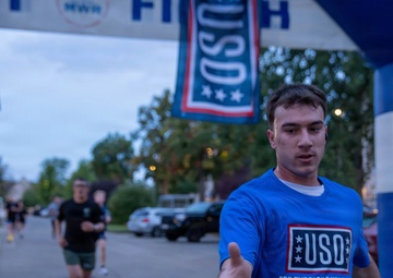 A runner wearing a blue USO t-shirt is crossing or approaching a finish line at what appears to be a charity or military-affiliated 5K/fun run event. He is reaching out to accept a water bottle being handed to him by a volunteer. The event is co-sponsored by USO and MWR (Morale, Welfare, and Recreation), suggesting a military installation setting.