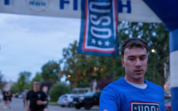 A runner wearing a blue USO t-shirt is crossing or approaching a finish line at what appears to be a charity or military-affiliated 5K/fun run event. He is reaching out to accept a water bottle being handed to him by a volunteer. The event is co-sponsored by USO and MWR (Morale, Welfare, and Recreation), suggesting a military installation setting.