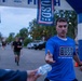 A runner wearing a blue USO t-shirt is crossing or approaching a finish line at what appears to be a charity or military-affiliated 5K/fun run event. He is reaching out to accept a water bottle being handed to him by a volunteer. The event is co-sponsored by USO and MWR (Morale, Welfare, and Recreation), suggesting a military installation setting.