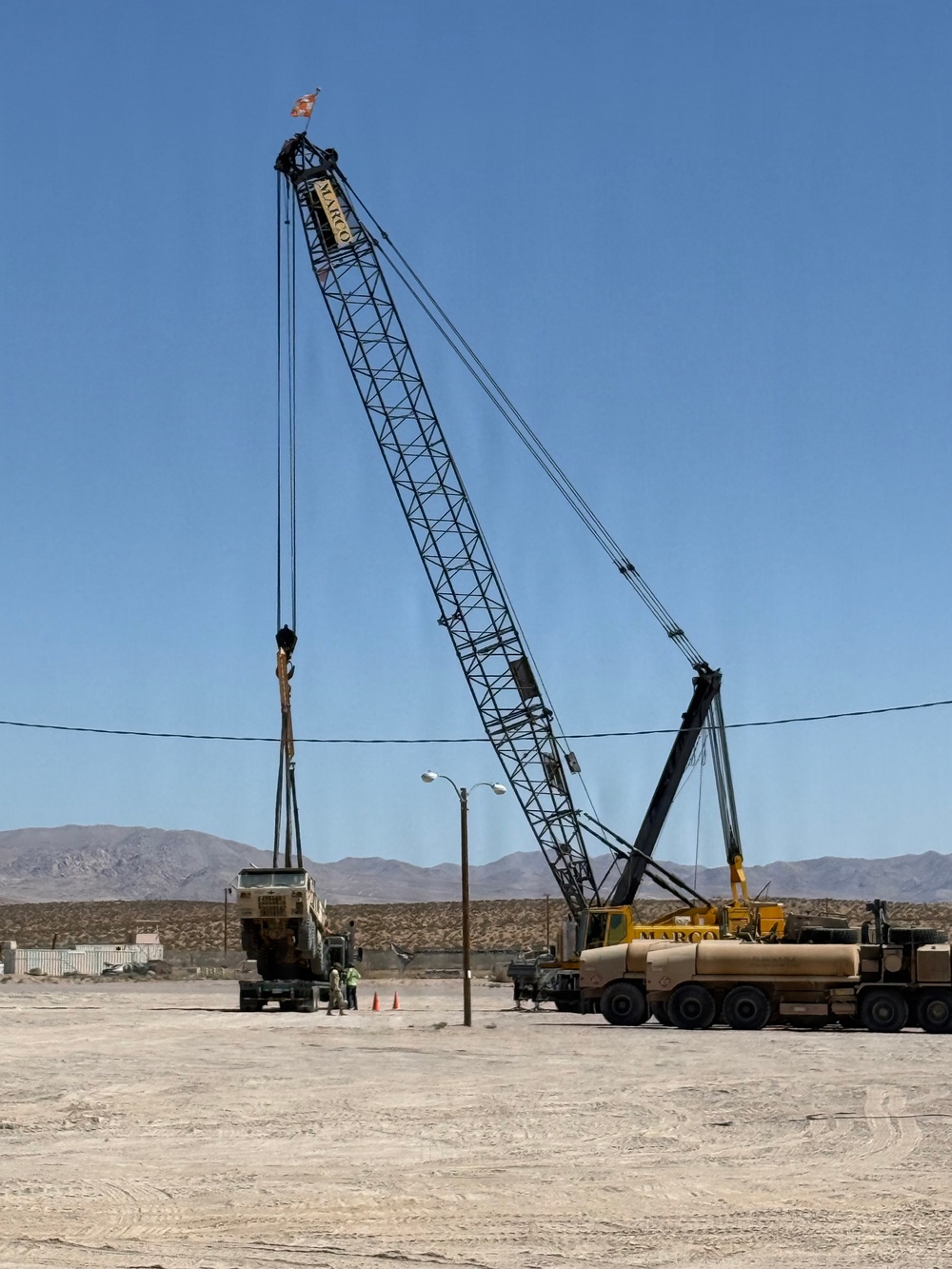 A large lattice boom crane branded 'MARCO' is operating in an arid desert environment, appearing to lift or position a heavy military-style vehicle. The scene takes place on a flat, sandy lot with mountains visible in the background under a clear blue sky. Safety cones and personnel are visible near the load.