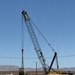 A large lattice boom crane branded 'MARCO' is operating in an arid desert environment, appearing to lift or position a heavy military-style vehicle. The scene takes place on a flat, sandy lot with mountains visible in the background under a clear blue sky. Safety cones and personnel are visible near the load.