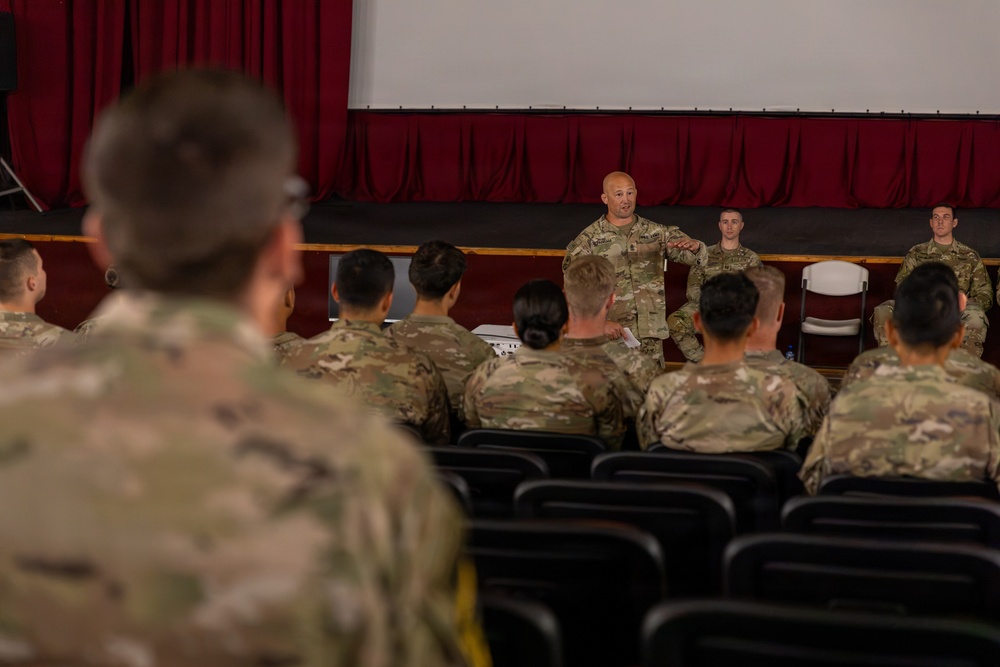 A military briefing or formation is taking place in what appears to be an auditorium or theater setting. One soldier stands at the front addressing a seated audience of uniformed personnel, while two additional soldiers stand on a raised stage behind him. The setting features red curtains, a projection screen, and rows of black auditorium seats.