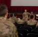 A military briefing or formation is taking place in what appears to be an auditorium or theater setting. One soldier stands at the front addressing a seated audience of uniformed personnel, while two additional soldiers stand on a raised stage behind him. The setting features red curtains, a projection screen, and rows of black auditorium seats.