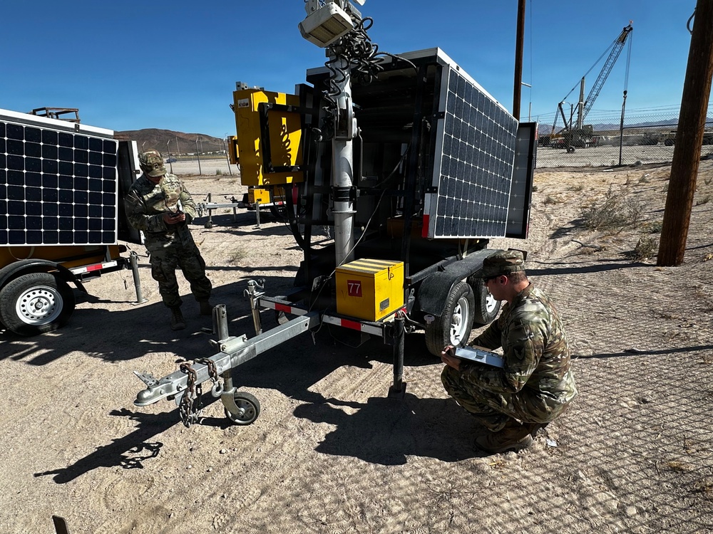 Two military personnel in camouflage uniforms are working with solar-powered mobile trailer units in an arid desert environment. One soldier is crouching near the trailer hitch area reviewing a clipboard or document, while another stands near a separate solar panel trailer in the background. The equipment appears to be mobile surveillance or communications towers mounted on trailers with solar panels.