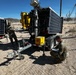 Two military personnel in camouflage uniforms are working with solar-powered mobile trailer units in an arid desert environment. One soldier is crouching near the trailer hitch area reviewing a clipboard or document, while another stands near a separate solar panel trailer in the background. The equipment appears to be mobile surveillance or communications towers mounted on trailers with solar panels.