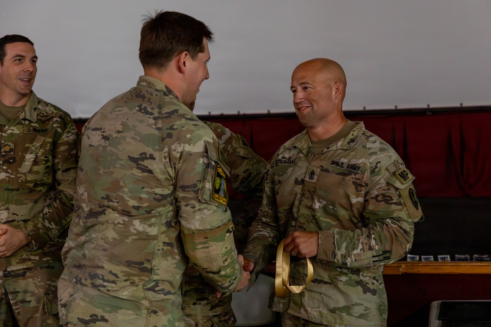 Two U.S. Army soldiers are engaged in a handshake during what appears to be a formal military ceremony or award presentation. The soldier on the right, wearing U.S. Army and MP (Military Police) insignia, is holding a gold ribbon or medal. A third soldier is visible smiling in the background.