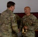 Two U.S. Army soldiers are engaged in a handshake during what appears to be a formal military ceremony or award presentation. The soldier on the right, wearing U.S. Army and MP (Military Police) insignia, is holding a gold ribbon or medal. A third soldier is visible smiling in the background.