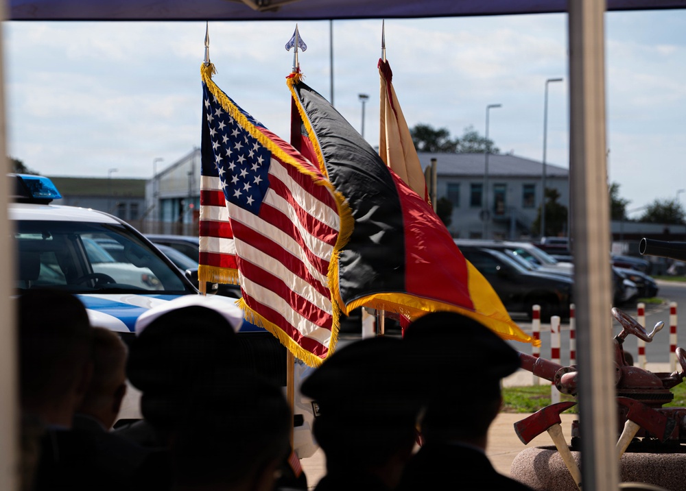An outdoor ceremony featuring a color guard presenting multiple flags, including the United States flag and what appears to be the German flag (black, red, and gold), suggesting a joint U.S.-German official event. Silhouetted figures wearing dark uniforms and caps, likely military or emergency services personnel, are visible in the foreground facing the flags. The setting appears to be a military base or official installation, with police/emergency vehicles visible in the background.