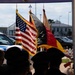 An outdoor ceremony featuring a color guard presenting multiple flags, including the United States flag and what appears to be the German flag (black, red, and gold), suggesting a joint U.S.-German official event. Silhouetted figures wearing dark uniforms and caps, likely military or emergency services personnel, are visible in the foreground facing the flags. The setting appears to be a military base or official installation, with police/emergency vehicles visible in the background.