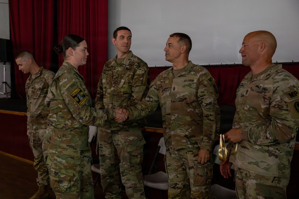 A U.S. Army military ceremony is taking place in what appears to be an auditorium or assembly hall. A female soldier is shaking hands with a male soldier identified by a 'SMITH' name tape, while other soldiers observe. The personnel wear Operational Camouflage Pattern (OCP) uniforms with 'MP' (Military Police) branch insignia visible on at least two individuals.