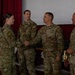 A U.S. Army military ceremony is taking place in what appears to be an auditorium or assembly hall. A female soldier is shaking hands with a male soldier identified by a 'SMITH' name tape, while other soldiers observe. The personnel wear Operational Camouflage Pattern (OCP) uniforms with 'MP' (Military Police) branch insignia visible on at least two individuals.