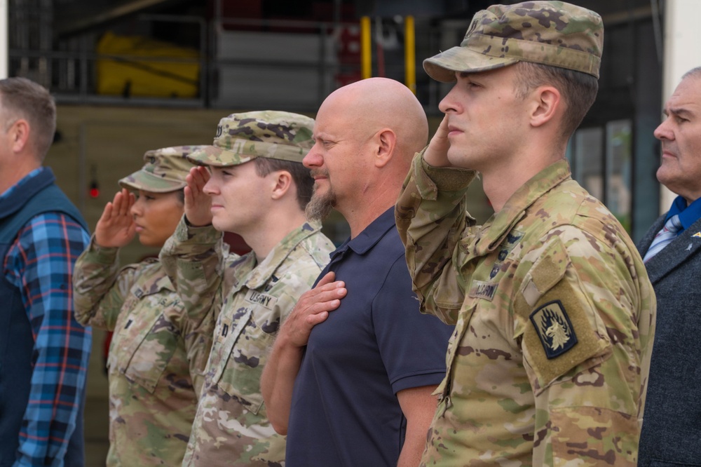 A group of U.S. Army soldiers in OCP (Operational Camouflage Pattern) uniforms are standing at attention and saluting during what appears to be a military ceremony, likely a national anthem or flag presentation. A civilian man in a dark polo shirt stands among them with his right hand placed over his heart. The setting appears to be an outdoor military installation with what looks like a fire station or vehicle bay in the background.