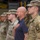 Katterbach Community and German Partners Commemorate 9/11 with Memorial Run and Ceremony A group of U.S. Army soldiers in OCP (Operational Camouflage Pattern) uniforms are standing at attention and saluting during what appears to be a military ceremony, likely a national anthem or flag presentation. A civilian man in a dark polo shirt stands among them with his right hand placed over his heart. The setting appears to be an outdoor military installation with what looks like a fire station or vehicle bay in the background.