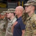 A group of U.S. Army soldiers in OCP (Operational Camouflage Pattern) uniforms are standing at attention and saluting during what appears to be a military ceremony, likely a national anthem or flag presentation. A civilian man in a dark polo shirt stands among them with his right hand placed over his heart. The setting appears to be an outdoor military installation with what looks like a fire station or vehicle bay in the background.