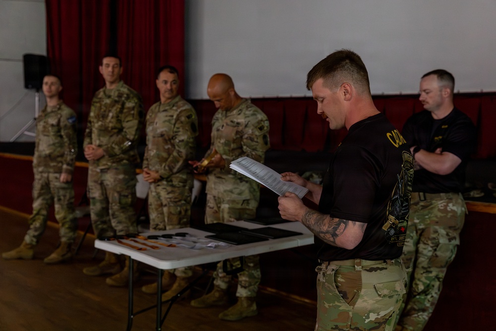 A military ceremony or briefing is taking place indoors, likely in a gymnasium or assembly hall. A soldier in a black t-shirt is reading from papers at a table while four uniformed soldiers in camouflage stand in a line behind him. Items including what appear to be awards or documents are laid out on a folding table in the foreground.