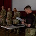 A military ceremony or briefing is taking place indoors, likely in a gymnasium or assembly hall. A soldier in a black t-shirt is reading from papers at a table while four uniformed soldiers in camouflage stand in a line behind him. Items including what appear to be awards or documents are laid out on a folding table in the foreground.