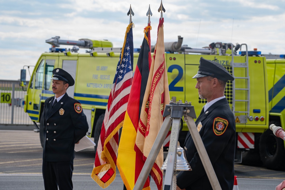 A formal ceremony is taking place at what appears to be a US Army Fire-Rescue station at Katterbach Airfield, Germany. Two firefighters in dress uniforms stand flanking a color guard display featuring the American flag, the German flag, and an Installation Management Command banner. A bright yellow-green fire rescue vehicle is prominently visible in the background.