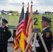 A formal ceremony is taking place at what appears to be a US Army Fire-Rescue station at Katterbach Airfield, Germany. Two firefighters in dress uniforms stand flanking a color guard display featuring the American flag, the German flag, and an Installation Management Command banner. A bright yellow-green fire rescue vehicle is prominently visible in the background.