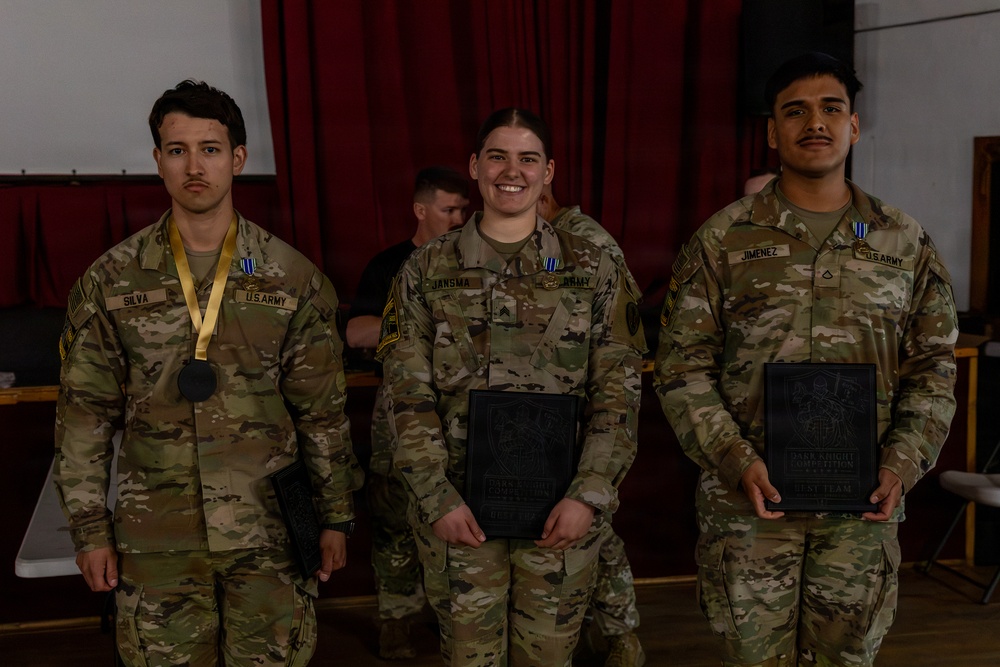 Three U.S. Army soldiers in OCP (Operational Camouflage Pattern) uniforms pose together at what appears to be an awards ceremony. The soldier on the left (Silva) wears a medal with a gold ribbon, while the soldiers in the center (Jansma) and right (Jimenez) hold dark plaques. The setting appears to be an indoor venue with red curtains in the background.