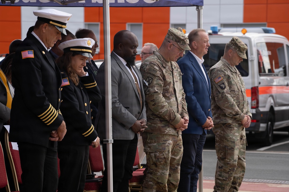 A formal outdoor ceremony featuring military personnel, civilians, and officials standing with heads bowed, likely in a moment of prayer or silence. The group includes U.S. Army soldiers in OCP camouflage uniforms, naval/fire department officers in dress uniforms with gold insignia, and civilians in business attire. The event appears to take place in a parking or staging area with emergency vehicles visible in the background.