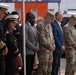 A formal outdoor ceremony featuring military personnel, civilians, and officials standing with heads bowed, likely in a moment of prayer or silence. The group includes U.S. Army soldiers in OCP camouflage uniforms, naval/fire department officers in dress uniforms with gold insignia, and civilians in business attire. The event appears to take place in a parking or staging area with emergency vehicles visible in the background.