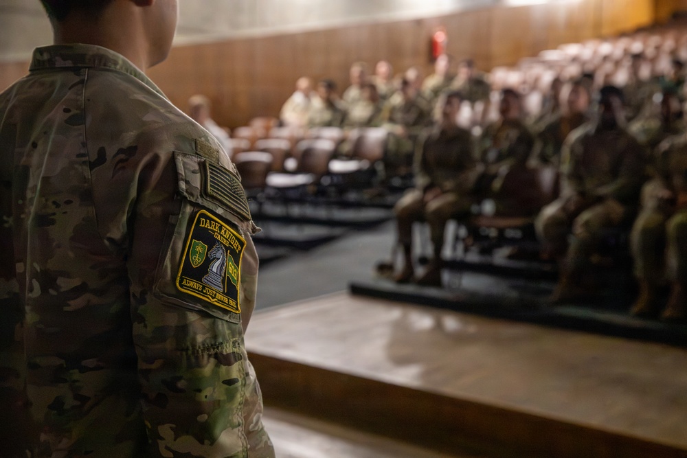 A military service member wearing OCP (Operational Camouflage Pattern) uniform stands at the front of an auditorium or briefing theater, facing a seated audience of fellow soldiers. The foreground figure displays a 'Dark Knight' unit patch on their left sleeve along with a US flag patch. The setting appears to be a formal military briefing, training session, or unit assembly.