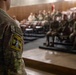 A military service member wearing OCP (Operational Camouflage Pattern) uniform stands at the front of an auditorium or briefing theater, facing a seated audience of fellow soldiers. The foreground figure displays a 'Dark Knight' unit patch on their left sleeve along with a US flag patch. The setting appears to be a formal military briefing, training session, or unit assembly.