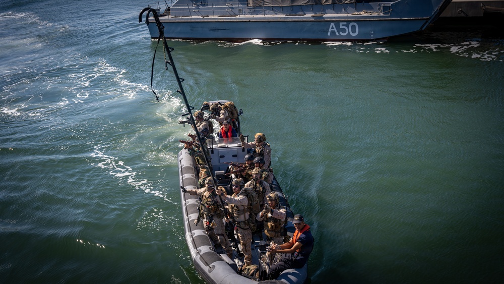 An aerial view shows a rigid inflatable boat (RIB) carrying approximately 10-12 armed military personnel in combat gear approaching a larger naval vessel marked 'A50'. The personnel appear to be conducting a maritime boarding or vessel approach exercise, with a grappling hook visible extending toward the larger ship. Two individuals wearing orange safety vests are visible among the group, likely serving as safety or boat crew personnel.