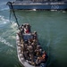An aerial view shows a rigid inflatable boat (RIB) carrying approximately 10-12 armed military personnel in combat gear approaching a larger naval vessel marked 'A50'. The personnel appear to be conducting a maritime boarding or vessel approach exercise, with a grappling hook visible extending toward the larger ship. Two individuals wearing orange safety vests are visible among the group, likely serving as safety or boat crew personnel.
