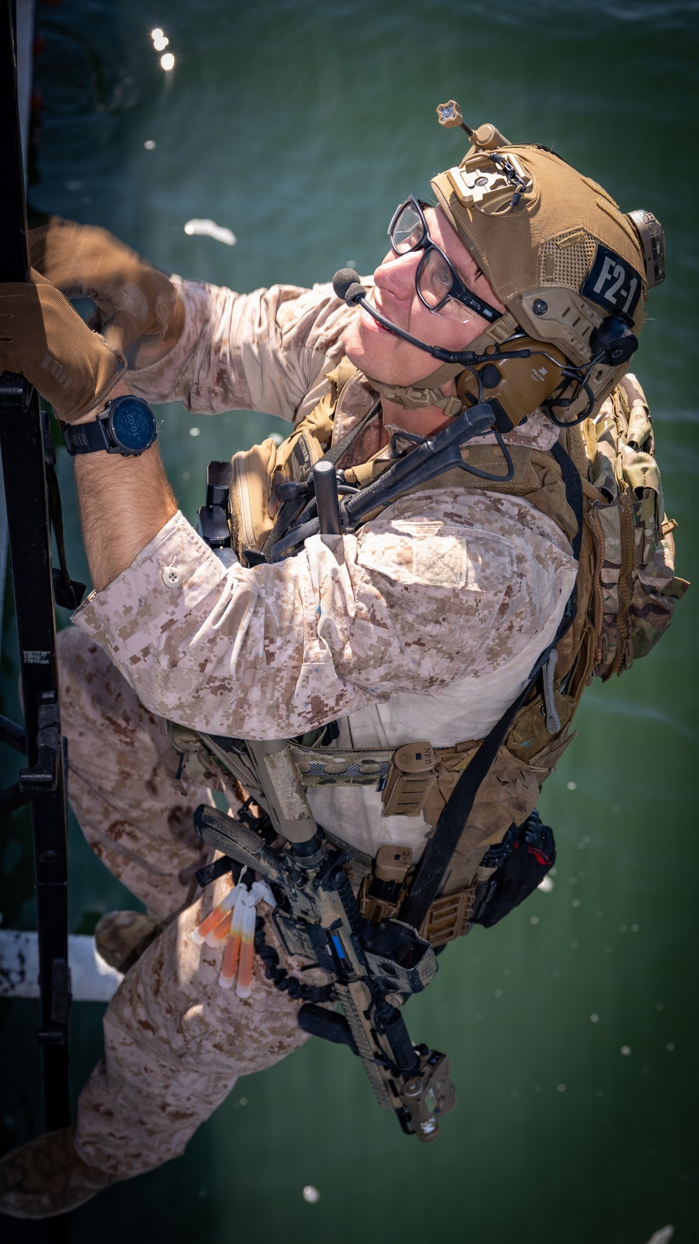A heavily equipped military operator in USMC MARPAT desert camouflage is climbing a ladder or rope structure over open water. The individual is wearing full tactical gear including a tan helmet with NVG mount, communications headset, body armor, and multiple pouches. The image is rotated approximately 90 degrees, giving the water a vertical background appearance.