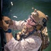 A heavily equipped military operator in USMC MARPAT desert camouflage is climbing a ladder or rope structure over open water. The individual is wearing full tactical gear including a tan helmet with NVG mount, communications headset, body armor, and multiple pouches. The image is rotated approximately 90 degrees, giving the water a vertical background appearance.
