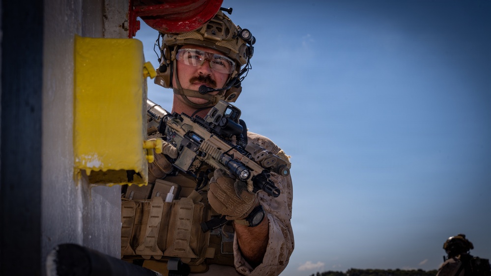 A heavily equipped military or special operations personnel is positioned against a wall, holding a tactical rifle in a ready position. The individual is wearing full combat gear including a tan/coyote brown plate carrier, tactical helmet with accessories, clear ballistic eyewear, and a communications headset. The image appears to capture a tactical training exercise or operational scenario.